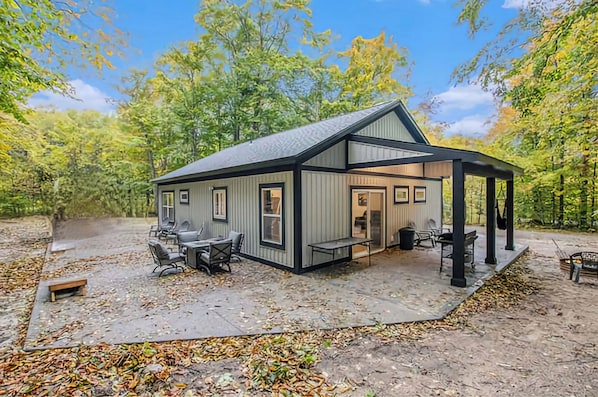 A small cabin with a covered patio area, outdoor seating, and a view of the surrounding forest.