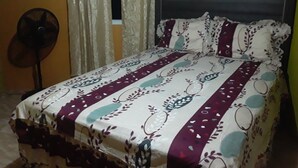 Un lit avec une couvre-lit à motifs et des oreillers, un ventilateur sur pied et des rideaux dans une pièce dont le mur est uni.