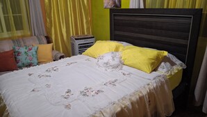 Un lit bien fait, avec un couvre-lit blanc et des oreillers jaunes, une tête de lit en bois et un petit climatiseur d’appoint dans un coin.