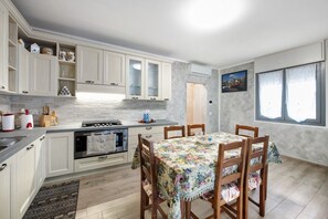 Une cuisine avec des meubles blancs, une table à manger recouverte d’une nappe à motifs floraux et une fenêtre donnant sur un paysage urbain.