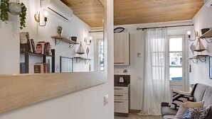 Una habitación con techo de madera, un área de cocina con armarios y hervidor, un sofá con cojines estampados, y un espejo grande que refleja la habitación.