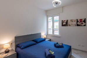 Un dormitorio con cama, mesita de noche, lámpara y una obra de arte en la pared.