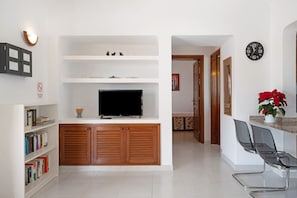 Una sala moderna con televisor de pantalla plana, armarios de madera, una estantería y una zona de estar con sillas.