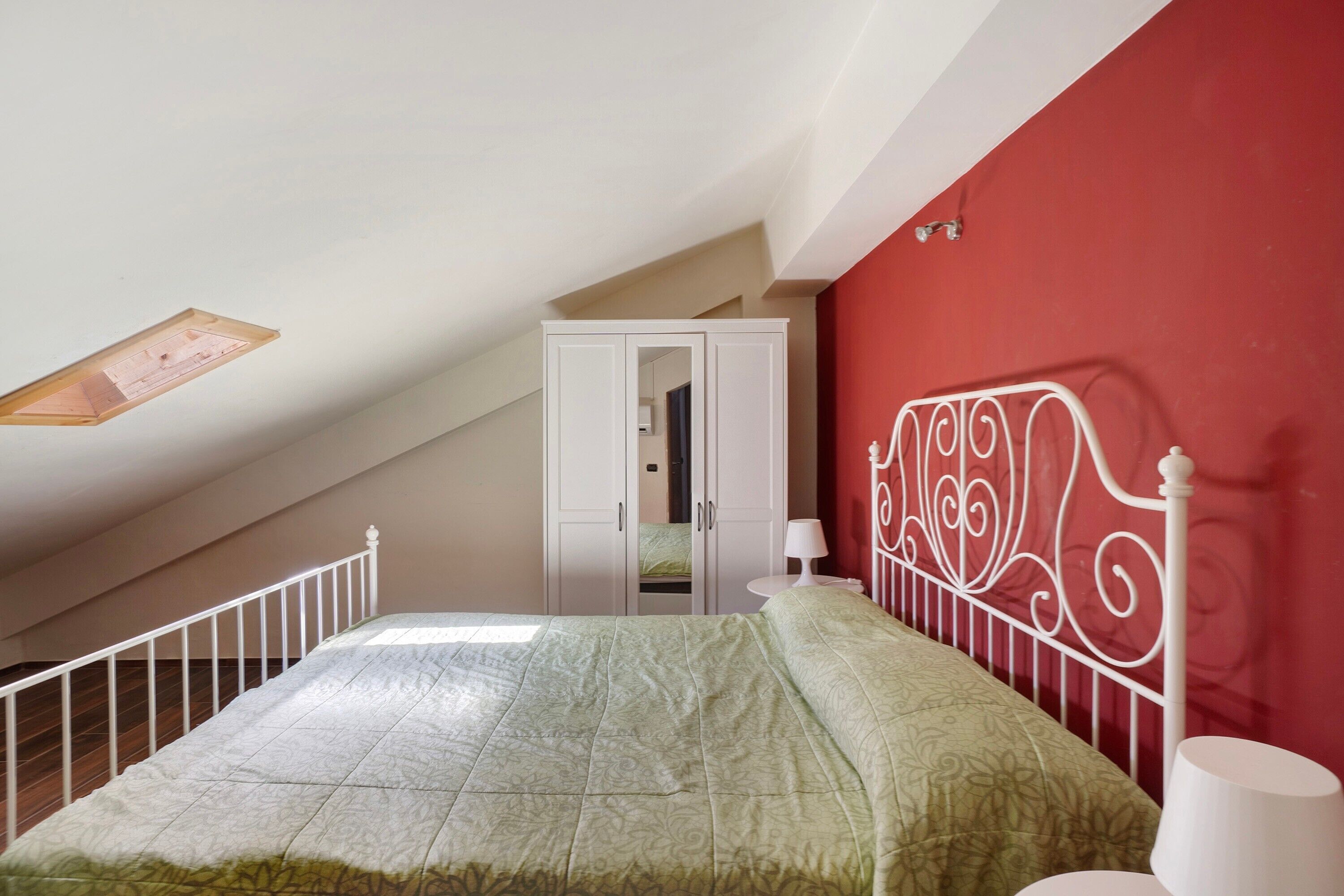 Une chambre avec un plafond en pente, un lit recouvert d’une couvre-lit à motifs verts, une tête de lit en métal blanche, une armoire blanche et une petite table de chevet avec une lampe.