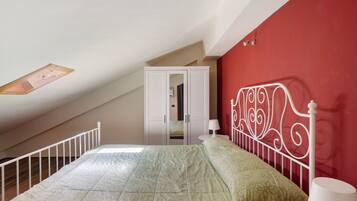 Une chambre avec un plafond en pente, un lit recouvert d’une couvre-lit à motifs verts, une tête de lit en métal blanche, une armoire blanche et une petite table de chevet avec une lampe.