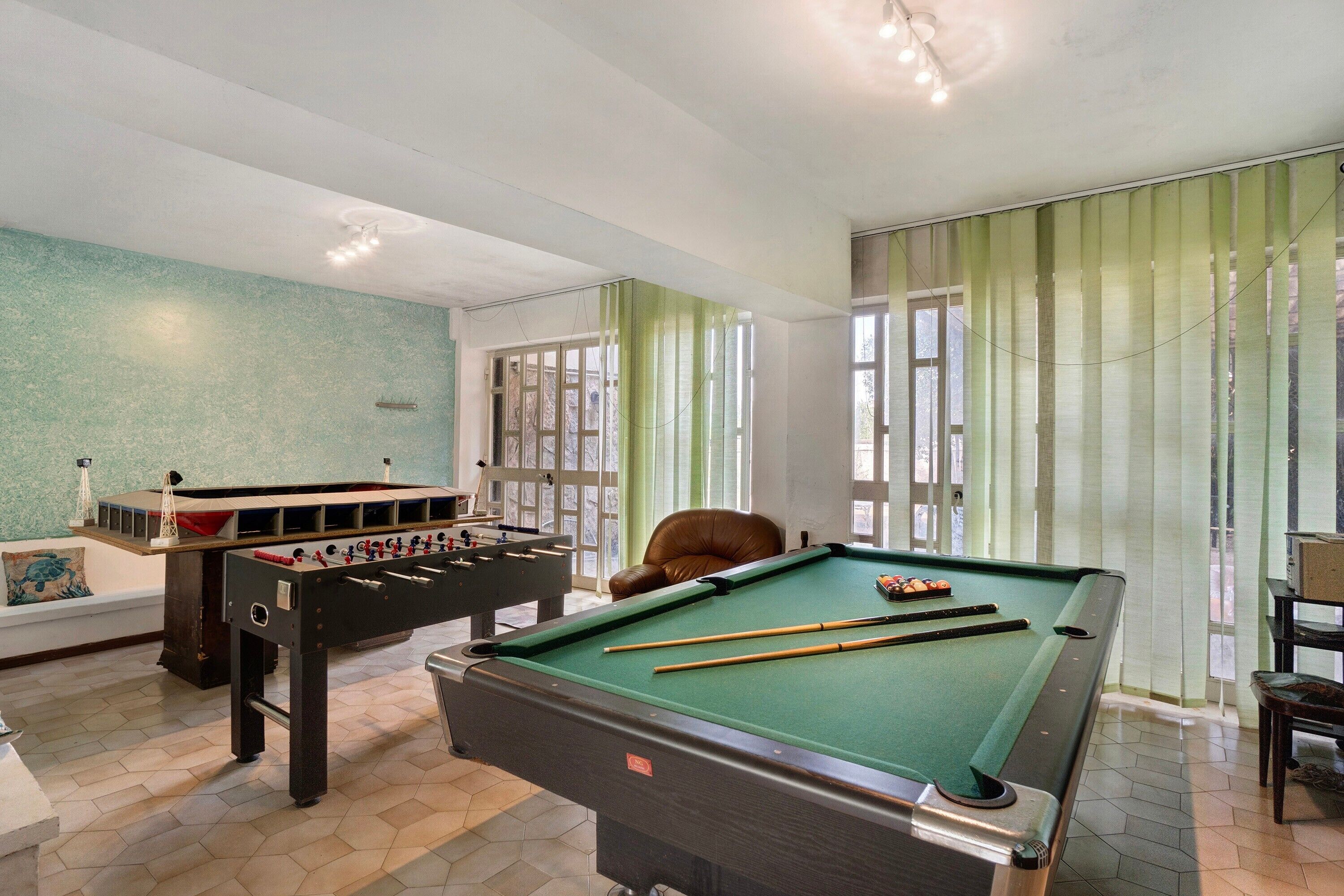 A pool table, foosball table, and a brown leather chair in a room with a green wall and large windows.