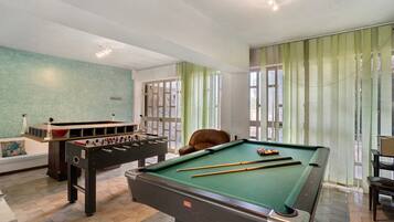 A pool table, foosball table, and a brown leather chair in a room with a green wall and large windows.