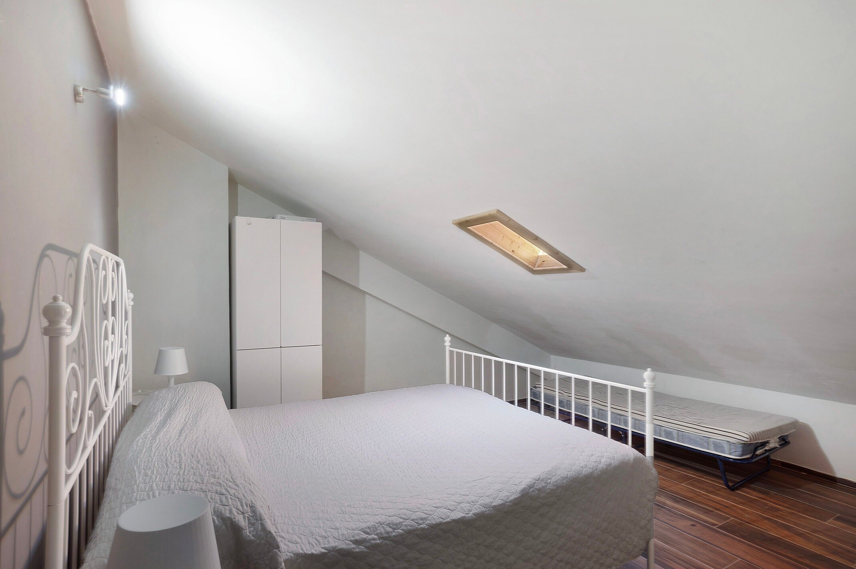 A bedroom with a sloped ceiling, a bed, a nightstand, a lamp, a mattress on the floor, and a small white cabinet.