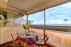 Une table recouverte d’une nappe colorée, un vase rempli de fleurs et un panier de pain, le tout sur un balcon donnant sur la mer.