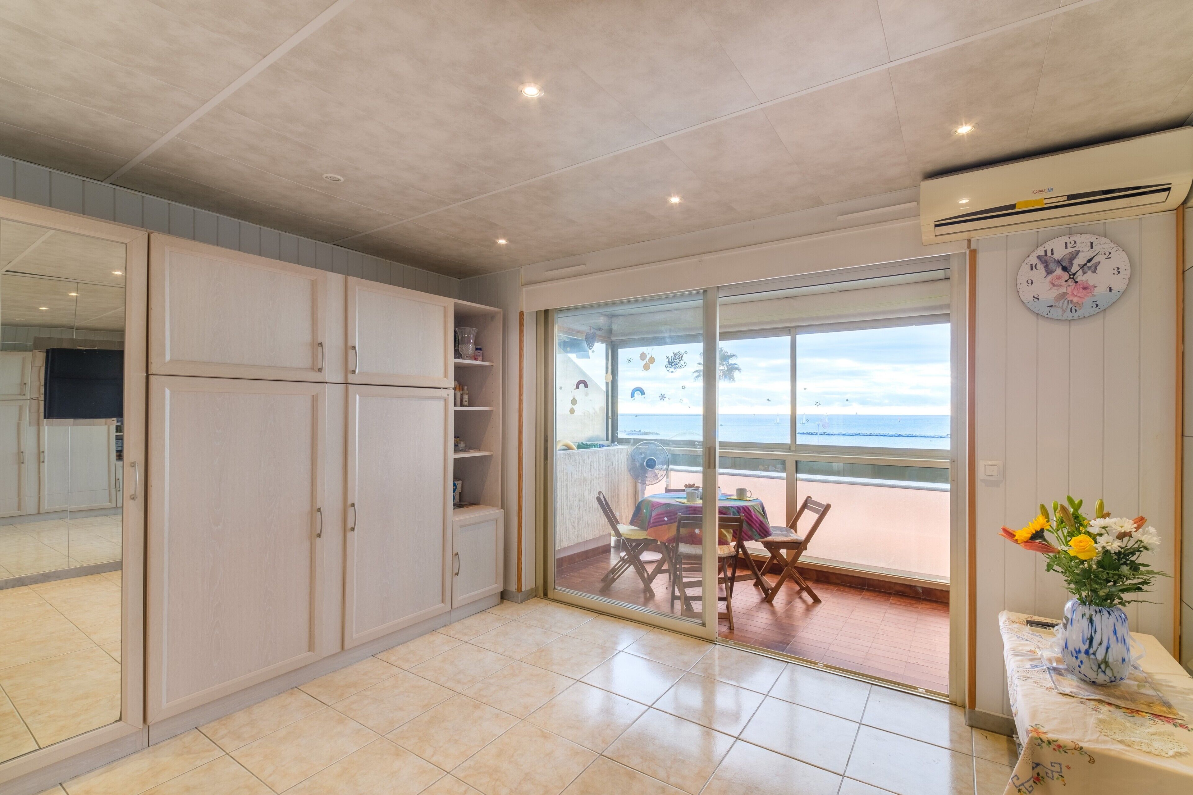 Ein Zimmer mit einer Glasschiebetür zu einem Balkon, einem Esstisch mit Stühlen, einer Uhr und einer Vase mit Blumen.