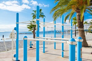 Des appareils de musculation en plein air, situés près d’une plage bordée de palmiers, sous un ciel dégagé.