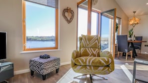Un salon spacieux doté d’une grande fenêtre offrant une vue sur la mer, d’un fauteuil confortable, d’un pouf à motifs zébrés et d’un coin repas agrémenté d’une plante et d’une bouteille.