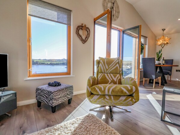 A spacious living room with a large window offering a view of the sea, a comfortable armchair, a zebra-patterned ottoman, and a dining area with a plant and a bottle.