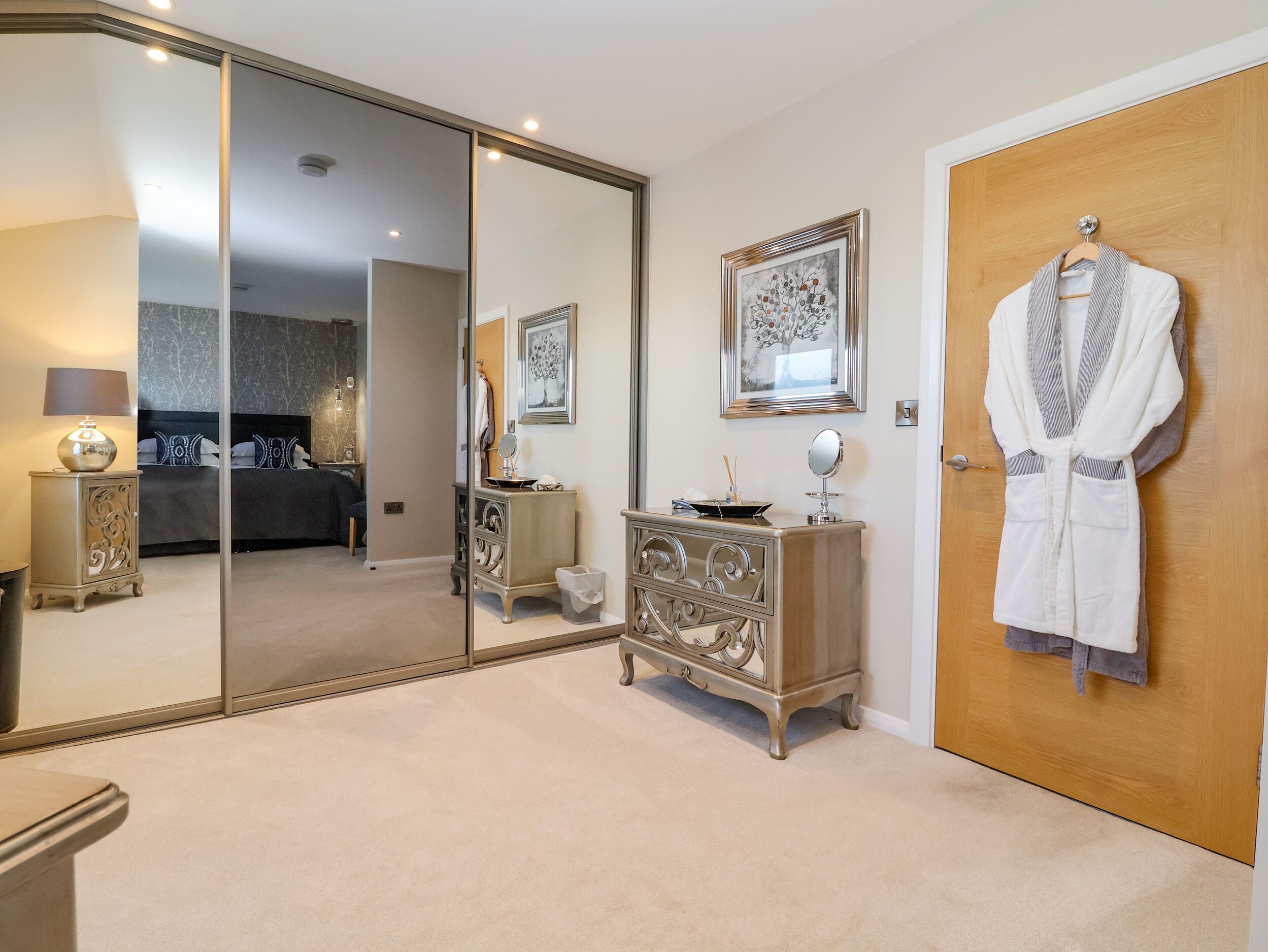 Une chambre à coucher avec un grand miroir, une armoire, une table de chevet avec une lampe et un lit doté d’une tête de lit sombre.