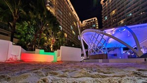 A sandy beach area with a modern building featuring a unique roof structure and illuminated by blue lighting. - Cozy 1BR cityview with Balcony and near Airport (Paranquae)
