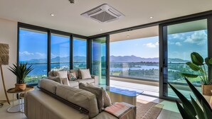 Una sala de estar moderna con un sofá grande, una mesa de centro y vistas a un paisaje urbano y montañas a través de amplios ventanales.