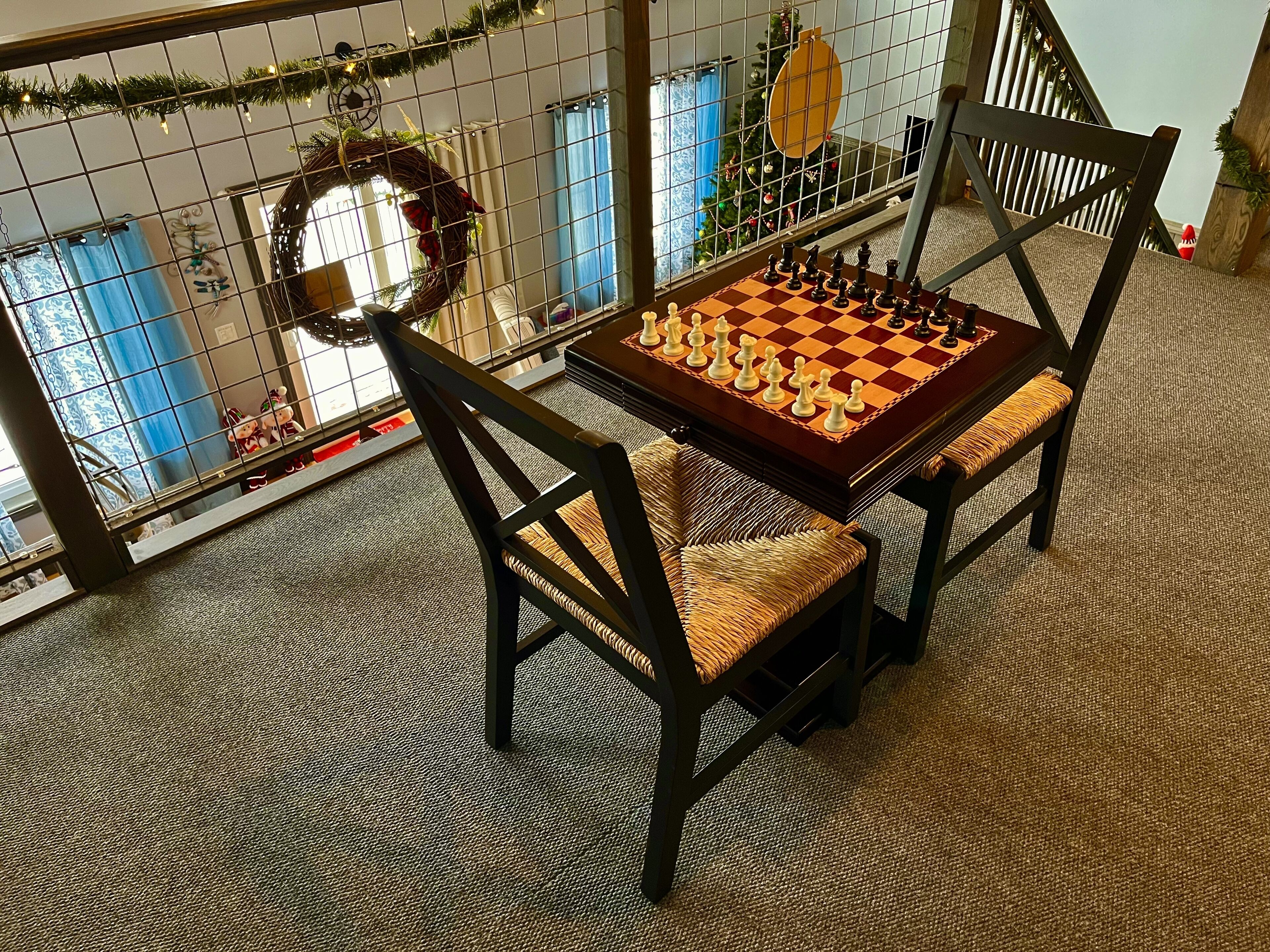 Un tablero de ajedrez sobre una mesa con sillas, un adorno navideño y vistas de un árbol de Navidad a través de una ventana.