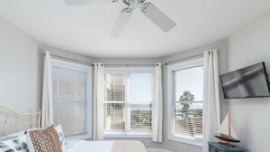 Een slaapkamer met een bed, een televisie, een klein tafeltje en grote ramen met jaloezieën.