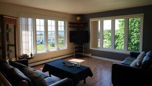 A living room with a sofa, a table, a TV, and large windows with a view of greenery. - Great Location - North Rustico Ocean View (North Rustico)