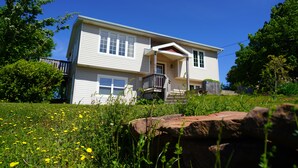 A two-story house with a front porch, a small deck, and a front yard with grass and wildflowers. - Great Location - North Rustico Ocean View (North Rustico)