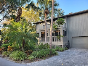 A two-story building with a balcony, surrounded by lush tropical plants and palm trees.