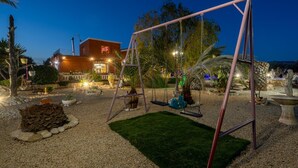 Un espacio exterior bien iluminado con un arenero, un pequeño jardín con césped y un edificio de fachada roja al fondo.