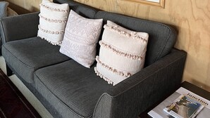 A grey sofa with three white pillows, a framed landscape painting, and a small table with a magazine. - Kākā Ridge Retreat Bush Hideaway (Tawharanui Peninsula)