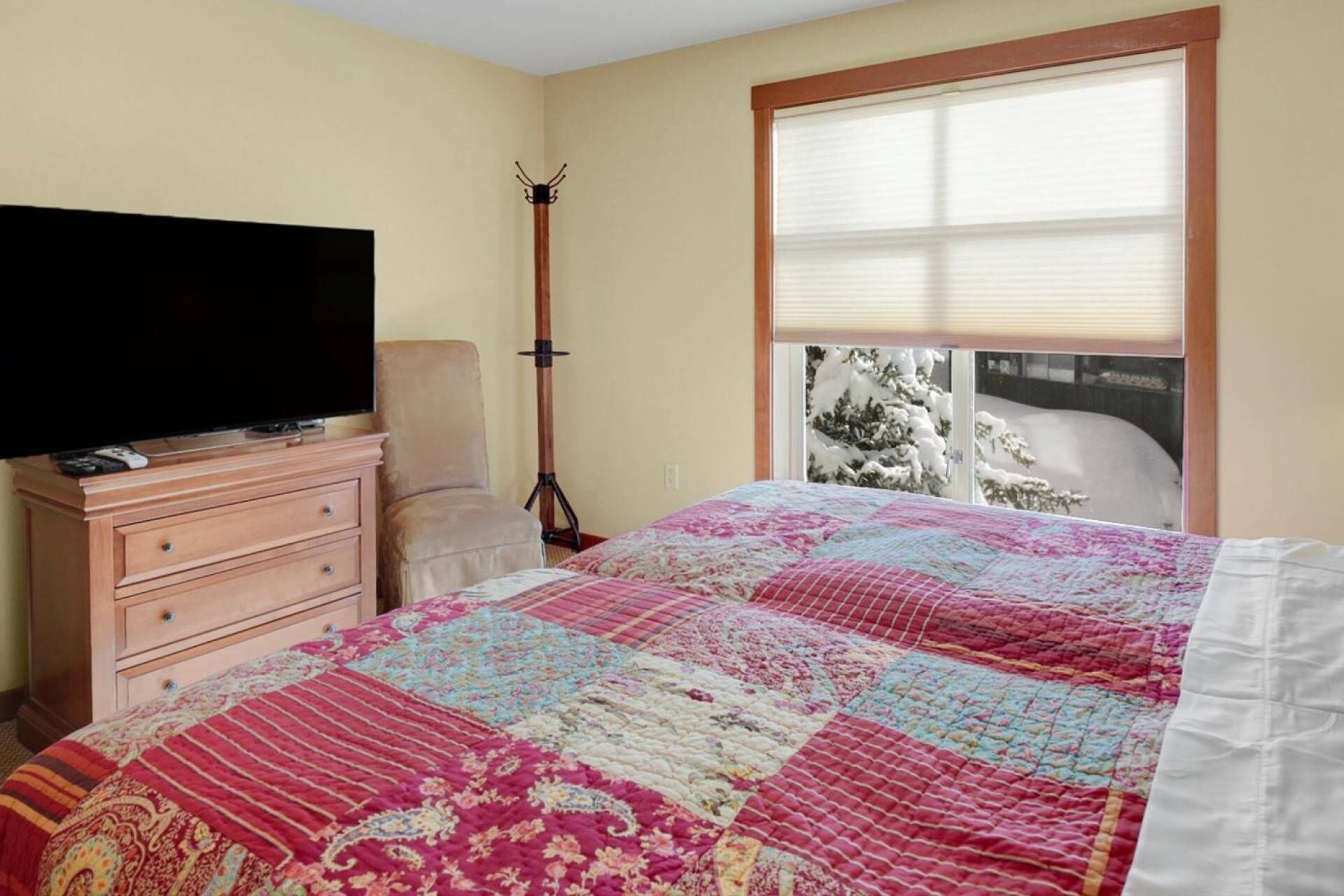 A bedroom with a bed, a dresser, a television, a chair, and a window with a view of snow-covered trees.