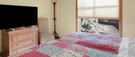 A bedroom with a bed, a dresser, a television, a chair, and a window with a view of snow-covered trees.