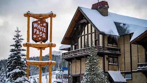 Ein hölzernes Schild mit der Aufschrift "WHITE PASS", ein Gebäude mit verschneitem Dach und verschneite Bäume im Hintergrund.