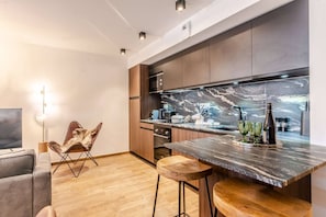 A modern kitchen with dark wood cabinets, a marble backsplash, and a built-in oven. There is a bar area with stools and a television in the adjacent living space. - Best location in Morzine, direct access to Avoriaz (MORZINE)