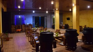 Um restaurante com mesas e cadeiras de madeira, um palco iluminado em azul e grandes vasos pretos no chão.
