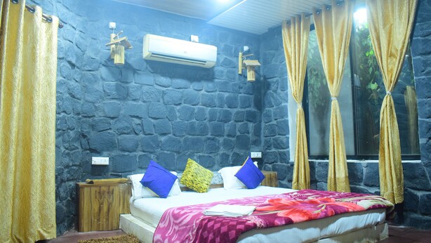 Quarto com parede de pedra, cama com manta rosa e vermelha, almofadas azuis e amarelas e cortinas douradas.