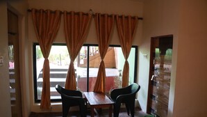 Quarto com mesa de madeira, duas cadeiras, janelas amplas com cortinas e um sapateiro encostado na parede.