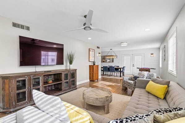 A spacious living room with a large flat-screen TV, a ceiling fan, and a dining area with a table and chairs.