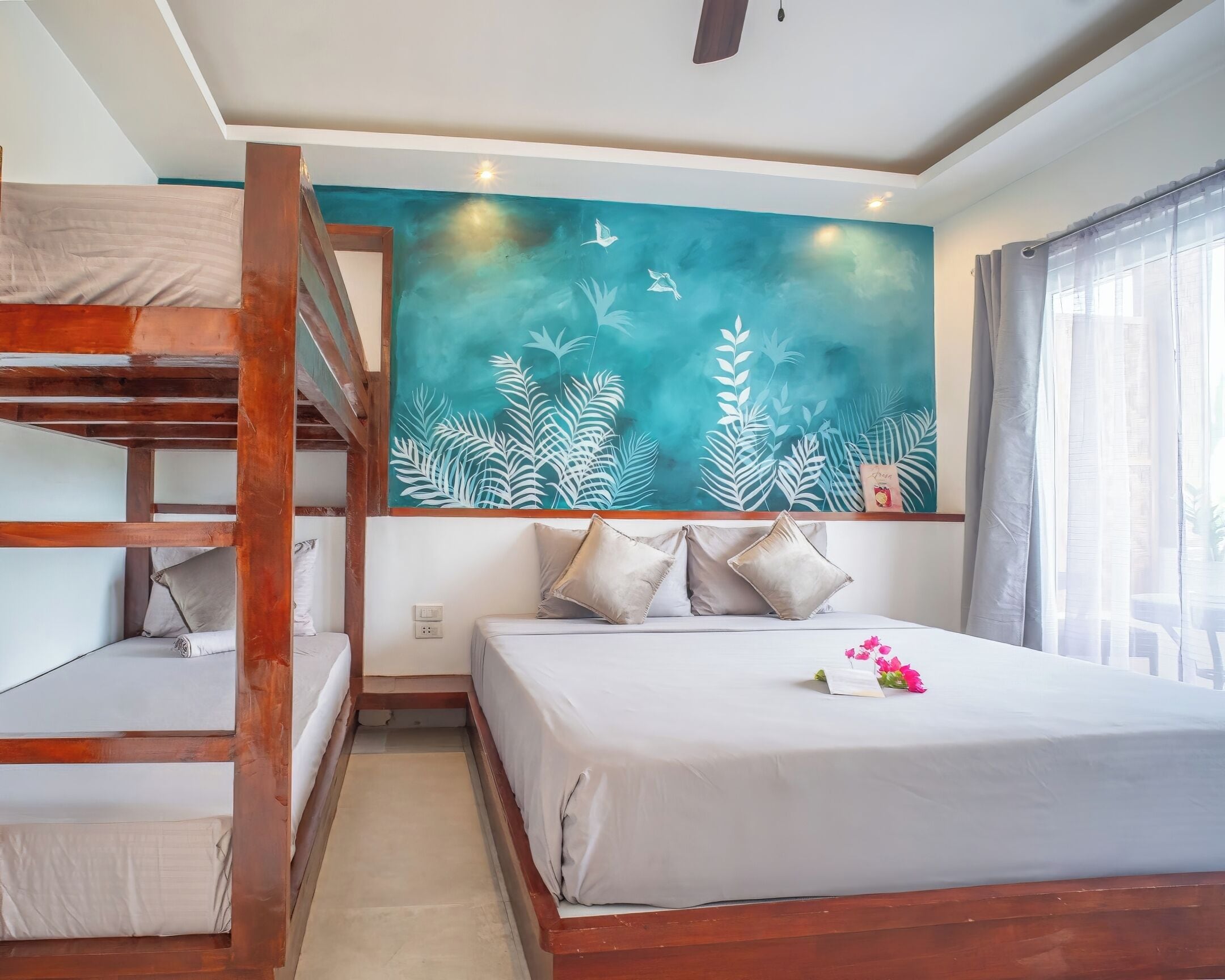 A hotel room with a bunk bed, a bed with pillows, a wall mural, and a window with curtains.