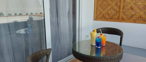A glass table with wicker chairs on a balcony with a view of a building and a woven wall panel.