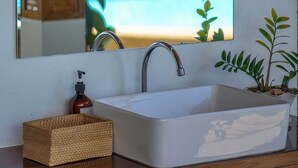 Un baño con lavamanos blanco, tocador de madera, espejo y vistas a una piscina y follaje.