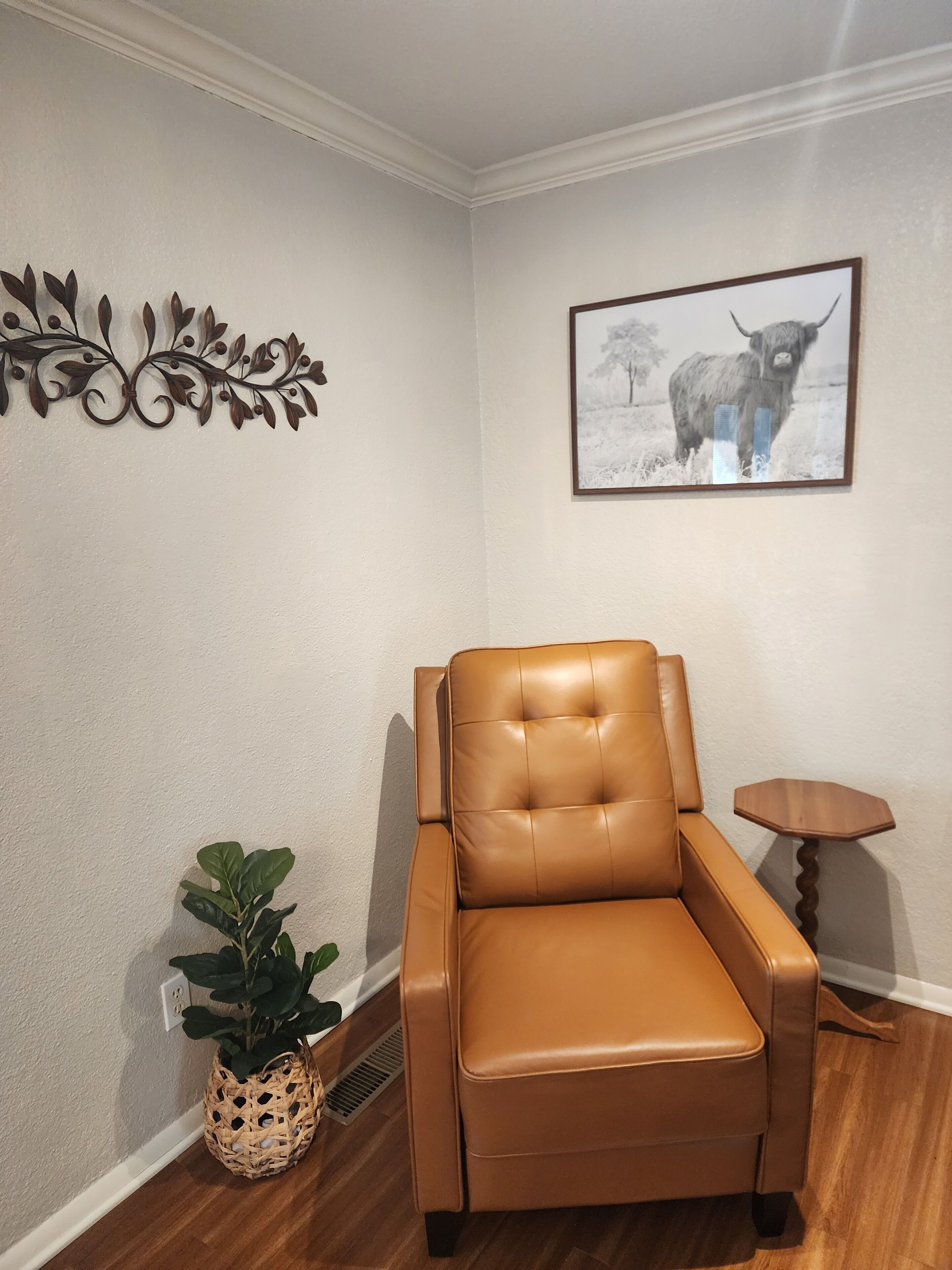 A tan leather chair in a corner of a room with a wooden floor, a small wooden side table, a wall-mounted decorative metal piece, and a framed picture of a yak.