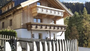 A two-story wooden chalet with a balcony, surrounded by a wooden fence and a forested backdrop. - Spacious apartment near the ski lift (Fendels)