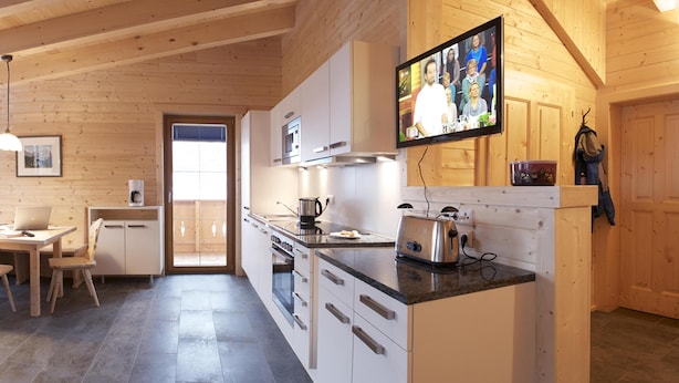 Une cuisine moderne avec des meubles en bois, un four encastré, une machine à café et un grille-pain. Un téléviseur fixé au mur diffuse une image d’un groupe de personnes.