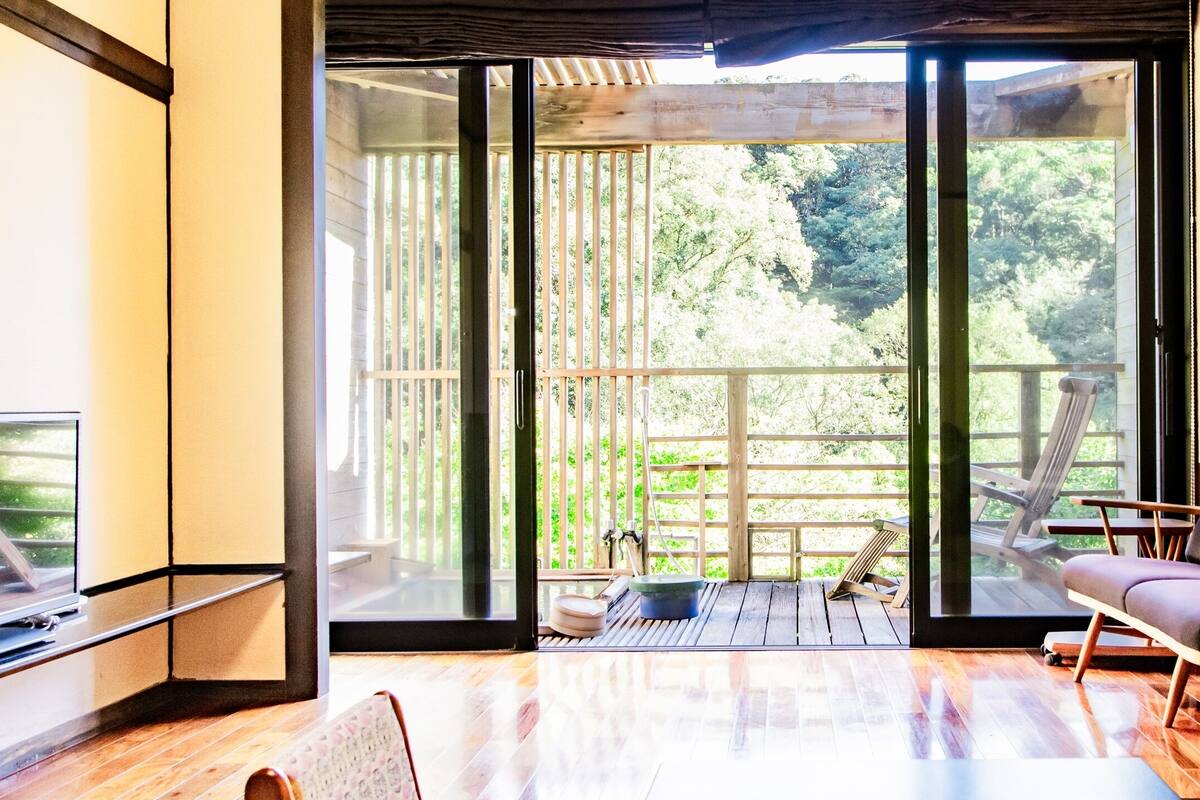 Une chambre moderne dotée d’une porte-fenêtre coulissante donnant sur une terrasse en bois, d’un fauteuil confortable et d’un téléviseur à écran plat.