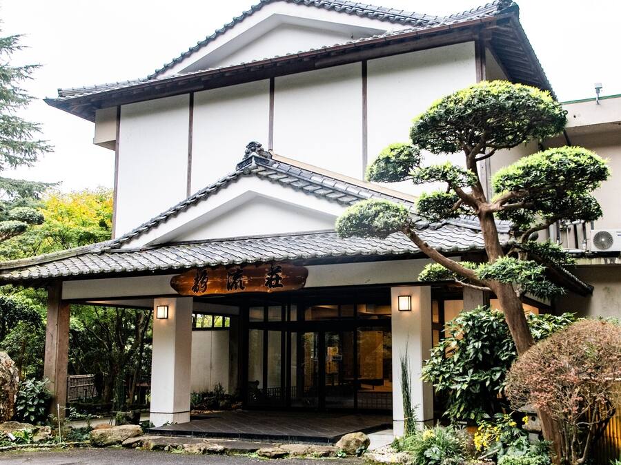 Een traditioneel Japans gebouw met een betegeld dak, omringd door verzorgde bomen en een pad van stenen dat naar de ingang leidt.