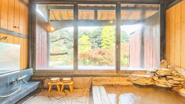 Un onsen japonés tradicional con una fuente termal de piedra, bancos de madera y vistas a follaje otoñal a través de amplias ventanas.