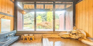 Ein traditionelles japanisches Onsen mit einem Steine-Thermalbad, Holzbänken und Blick auf herbstliche Laubfärbung durch große Fenster.