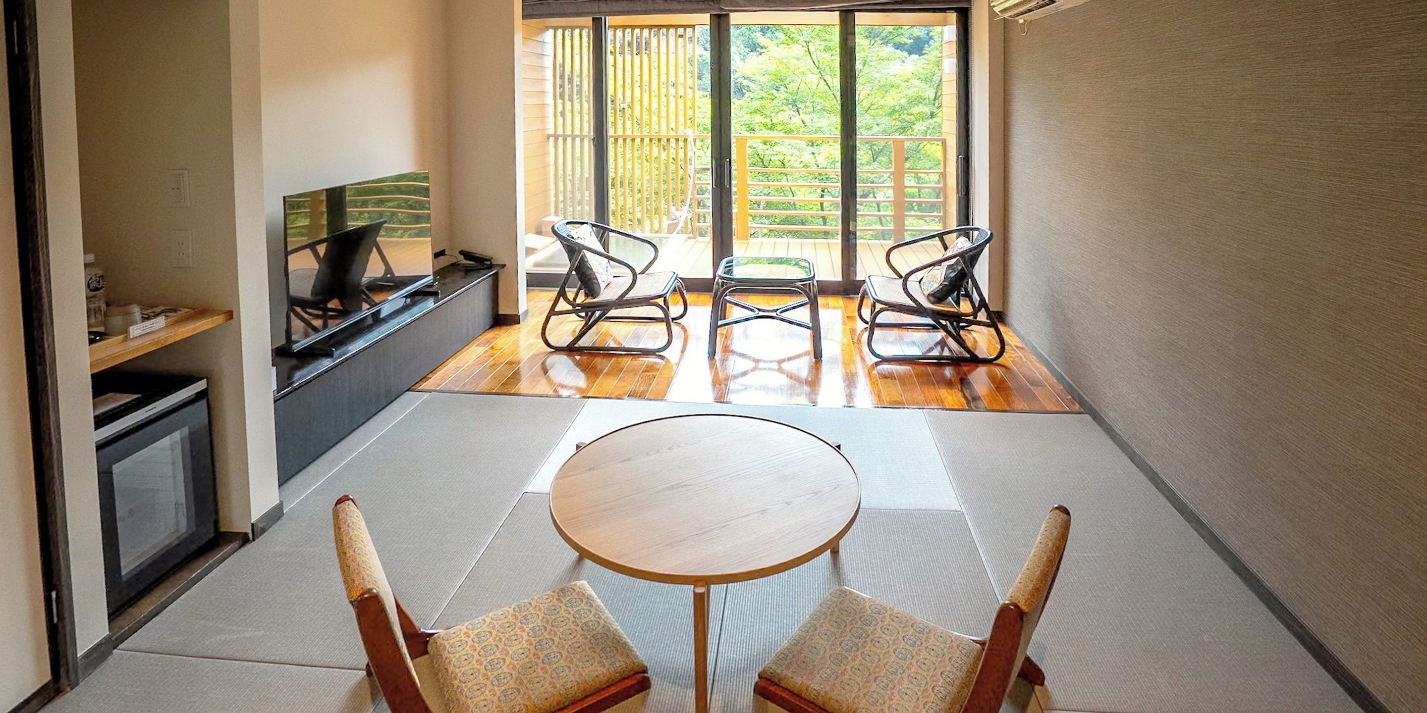 Quarto com piso de tatame, uma mesa baixa, duas cadeiras, uma televisão e uma porta de vidro deslizante que dá para uma varanda com vista para o verde.