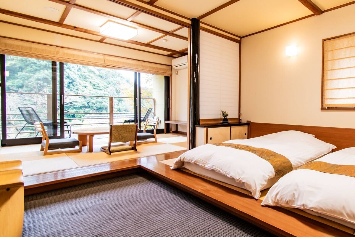 Une pièce de style japonais traditionnel, avec un sol recouvert de tatamis, un lit bas en bois avec une literie blanche, un paravent coulissant de type shoji et une vue sur une zone boisée.