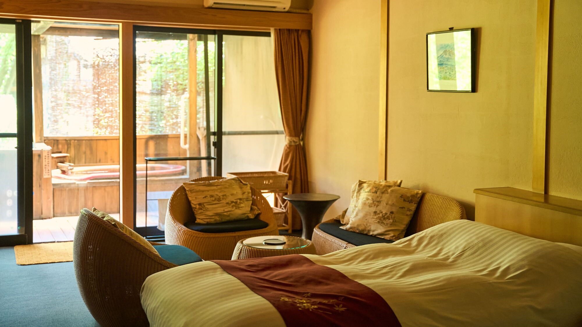 A hotel room with a bed, a chair, a small table, and a view through a sliding door to an outdoor area.