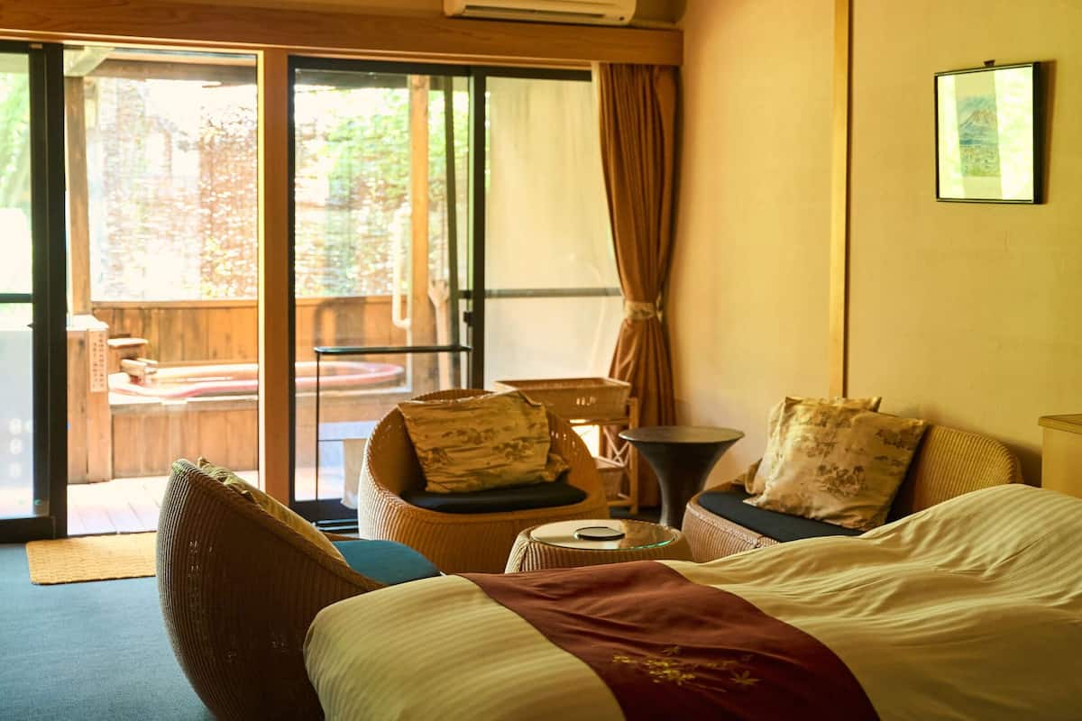 A hotel room with a bed, a chair, a small table, and a view through a sliding door to an outdoor area.