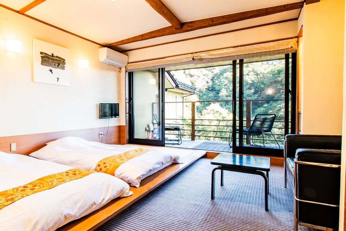 Une chambre d’hôtel avec deux lits, une petite table, une chaise et une grande fenêtre donnant sur l’extérieur.